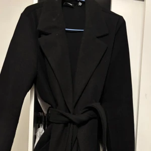 Svart kappa med bälte och krage - Stilren svart kappa med bred krage och knytbälte i midjan. Långärmad modell med klassisk siluett, perfekt för dig som gillar en clean och tidlös look. Materialet känns mjukt och ger en snygg drapering över axlarna.