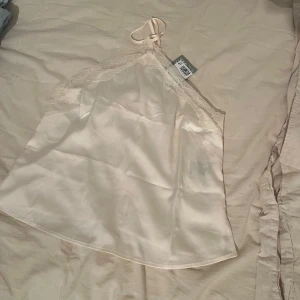 Champagnefärgad linnetopp med spets - Snygg champagnefärgad linnetopp i satinliknande material med tunna axelband och vacker spetsdetalj längs urringningen. Perfekt för lager på lager eller som statement-piece. Lätt och luftig känsla, passar till många olika stilar.