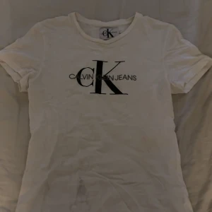 Vit Calvin Klein t-shirt - Vit t-shirt från Calvin Klein Jeans med klassisk svart CK-logga framtill.  Super fin och skön. Tyvärr har den fått en fläck som inte går bort i tvätten, tänkte att jag lägger ut den ändå då någon kanske vill klippa av den och ha som magtröja eller något liknande🩷