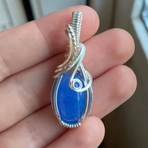 Silverberlock med blå sten - Unikt berlock till halsband med en oval, intensivt blå sten infattad i silverfärgad tråd. Stenen är slät och omges av konstnärligt tvinnad metalltråd som ger en cool, bohemisk vibe. Perfekt för dig som vill sticka ut med ett personligt smycke.