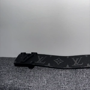 Louis Vuitton svart monogram bälte - Snyggt svart bälte från Louis Vuitton med klassiskt monogrammönster och mattsvart LV-spänne. Utsidan är svart med diskreta gråa loggor och insidan är ljusbrun med kontrastsömmar. Perfekt accessoar för att lyfta din outfit.