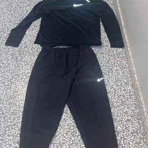 Svart långärmad Nike Dri-FIT träningströja - Svart långärmad träningströja från Nike med Dri-FIT-teknologi. Tröjan har en klassisk passform, rund hals och reflekterande Nike-logga på bröstet. Perfekt för träning och sportiga aktiviteter, tillverkad i lätt och snabbtorkande syntetmaterial.