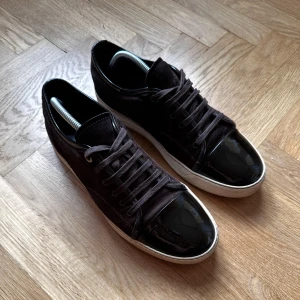 Bruna sneakers med lackad tå - Snygga bruna sneakers med ovandel i mocka och cool lackad svart tå. Skorna har vita sulor och klassisk snörning. Perfekta för dig som vill ha en stilren men ändå unik look till vardags.