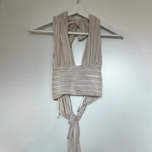 Champagnefärgad omlottopp från Gina Tricot XS - Supersnygg omlottopp i champagnefärgat glansigt tyg från Gina Tricot. Toppen har breda axelband, knyt i nacken och midjan samt plisserad struktur.