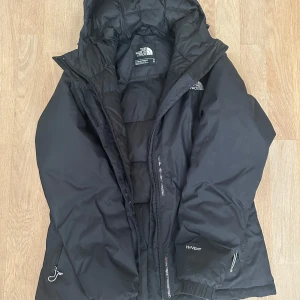 Svart dunjacka från The North Face M - Svart dunjacka från The North Face med huva och dragkedja. Jackan har quiltad design, elastiska muddar och snörning i nederkant. Perfekt för kalla dagar och har klassisk logga på bröstet. Snygg och funktionell för vintergarderoben. Den har ett hål som ni ser på sista bilden❤️