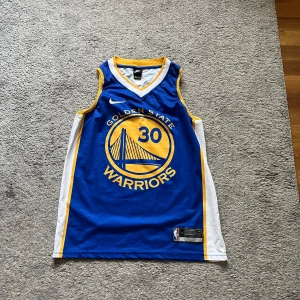 Golden State Warriors Steph Curry jearsey  - Golden State Warriors basketlinne från Nike med nummer 30 och Curry på ryggen. Linnet är blått med gula och vita detaljer, har ärmlös design och är gjort i lätt polyester. Perfekt för dig som älskar basket och vill representera Steph Curry.