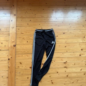 Svarta Adidas mjukisbyxor  - Svarta mjukisbyxor från Adidas med klassiska vita ränder längs sidorna och logga på låret. Byxorna har dragsko i midjan och är tillverkade i mjuk polyester, perfekta för chill eller träning. Snygg och sportig look som alltid funkar.
