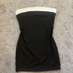 Svart axelbandslös kortklänning - Snygg svart axelbandslös kortklänning med vit kant upptill. Klänningen har en enkel och stilren design som sitter tight och markerar figuren. Perfekt för dig som gillar minimalistisk och klassisk stil.