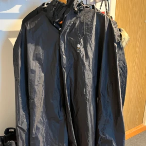 Svart vindjacka med huva från 2117 - Svart vindjacka från 2117 med huva och minimalistisk design. Jackan har dragkedja framtill och liten logga på bröstet. Perfekt för blåsiga dagar och enkel att slänga på sig över en hoodie.