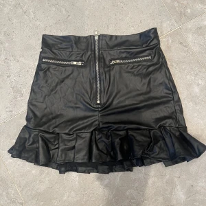 Svart kort kjol i skinnimitation - Snygg svart kort kjol i skinnimitation med volangkant nertill. Kjolen har två dragkedjor framtill och en stor dragkedja mitt bak för en edgy look. Perfekt för dig som vill sticka ut med en cool och trendig stil.