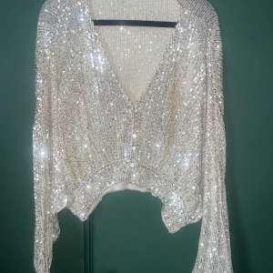 Glittrig topp från Pull&Bear - Superfin glittrig topp från Pull&Bear i guld med massor av paljetter. Toppen har v-ringning, knappar framtill och långa ärmar. Perfekt för dig som vill sticka ut och glänsa lite extra. Sitter snyggt och har en lite croppad passform. Använd 2 gånger