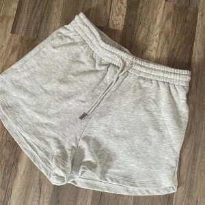 Ljusgrå sweatshorts från H&M - Superbekväma ljusgrå sweatshorts från H&M med elastisk midja och snörning. Tillverkade i en mjuk bomullsblandning med polyester och viskos. Perfekta för chill dagar eller träning. Klassisk loose fit och enkel design utan mönster. HELT OANVÄNDA