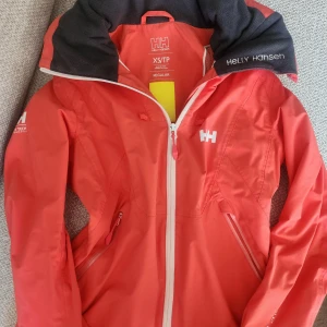 Röd vindjacka Helly Hansen XS - Snygg röd vindjacka från Helly Hansen i storlek XS. Jackan har vit dragkedja, Helly Tech Performance-tryck på ärmen och fleecefodrad krage. Perfekt för blåsiga dagar och outdoor-äventyr. Praktiska fickor med dragkedja och coola reflexdetaljer på ryggen.