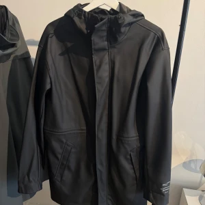 Svart regnjacka från Pull&Bear - Svart regnjacka från Pull&Bear Urban Theory med huva och dragkedja framtill. Jackan har två snedställda fickor och tryckt text på ärmen. Snygg minimalistisk design som funkar till alla outfits.