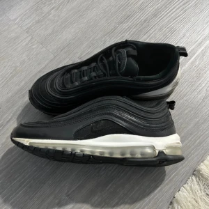 Nike Air Max 97 svart/vit strl 41 - Säljer ett par Nike Air Max 97 i svart med vita detaljer och reflekterande ränder. Skorna har en ikonisk vågig design, synlig Air-sula och snörning. Ovandelen är i mesh och skinn för en sportig look. Perfekta för dig som gillar streetwear och sneakers med attityd.