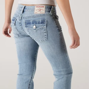 True Religion ljusblå bootcut jeans - Säljer ett par ljusblå jeans från True Religion med ikoniska bakfickor och snygga kontrastsömmar. Jeansen har bootcut-modell och låg midja, tillverkade i mjukt denim med klassisk knappstängning och bälteshällor. Perfekta för en avslappnad och trendig look.