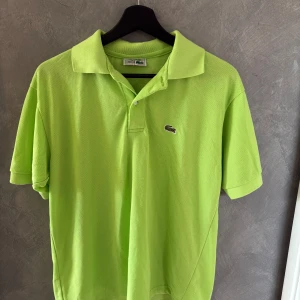 Grön pikétröja från Lacoste - Snygg limegrön pikétröja från Lacoste med klassisk krage och korta ärmar. Tröjan har den ikoniska krokodilloggan broderad på bröstet och är tillverkad i bomull. Perfekt för dig som vill sticka ut med en fräsch färg i sommar. (Liten i storleken)