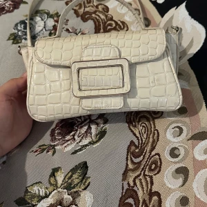 Vit croco handväska med stor spänne - Snygg vit handväska i croco-mönstrat syntetmaterial med en stor dekorativ spänne framtill. Väskan har både handtag och axelrem i silverkedja, perfekt för att lyfta din outfit. Stilren och trendig design med svart innerfoder och praktiskt fack.