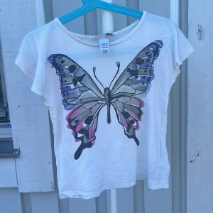 Vit topp med fjäril från H&M - Supersöt vit topp från H&M med en stor fjäril i lila, blått och rosa på framsidan. T-shirten har korta ärmar och rund halsringning. Perfekt för dig som gillar färgglada tryck och vill sticka ut lite extra.