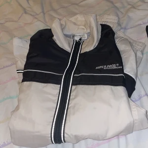 Svart och vit vindjacka Super Pros XL plus tillhörande byxor - Snygg svart och vit vindjacka från Super Pros i storlek XL. Jackan har dragkedja med reflexdetaljer, sportig design och meshfoder för extra komfort. Perfekt för dig som gillar en clean och stilren look.Plus vind byxor.