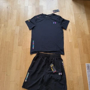 Svart Under Armour t-shirt i polyester - Svart t-shirt från Under Armour med färgglada loggor och detaljer. Tillverkad i lätt och ventilerande polyester, perfekt för träning. Klassisk rund hals och korta ärmar. Snygg och sportig vibe med regnbågsfärgade tryck på bröstet och axeln.