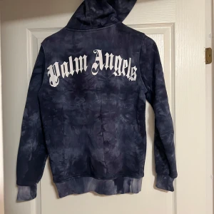 Palm Angels tie-dye hoodie marinblå - Snygg marinblå tie-dye hoodie från Palm Angels med stor vit logga på ryggen och mindre logga fram. Klassisk känguruficka, huva och ribbade muddar. Perfekt för dig som vill sticka ut med streetwear-vibe. OBS! Saknar snören i luvan!