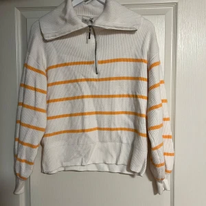 Randig vit och orange tröja från Y.A.S - Vit ribbstickad tröja från Y.A.S med orangea ränder och half zip-dragkedja vid kragen. Tröjan har lång ärm, hög krage och en relaxed fit. Perfekt för dig som gillar en sportig och chill stil med lite färg.
