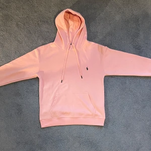 Rosa hoodie från Polo Ralph Lauren - Säljer en ljusrosa hoodie från Polo Ralph Lauren. Klassisk modell med huva, snörning och magficka. Liten broderad logga på bröstet. Perfekt för dig som gillar stilrena och bekväma plagg.