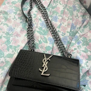 Svart YSL axelväska i skinn - Säljer en svart axelväska från YSL med croco-mönstrat skinn och silverfärgad kedja. Väskan har en stor YSL-logga framtill och stilren, boxig form. Perfekt för dig som vill ha en lyxig och edgy accessoar med plats för det viktigaste.
