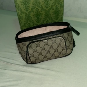 Gucci beige axelväska med GG-mönster - Säljer en snygg beige axelväska från Gucci med klassiskt GG-mönster och svarta skinn-detaljer. Väskan har justerbar rem, dragkedja upptill och ett extra fack framtill. Perfekt för dig som vill ha en stilren och ikonisk accessoar.