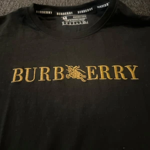  Burberry t-shirt - Svart t-shirt från Burberry i mjuk bomull med guldig broderad logga på bröstet och ärmen. Klassisk rund hals och korta ärmar. Snygg och stilren design som sticker ut med exklusiv känsla.