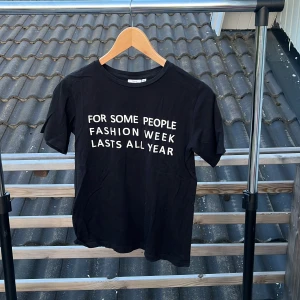 Svart t-shirt med text från KappAhl - Svart t-shirt från KappAhl i bomull med vit text på framsidan: 'FOR SOME PEOPLE FASHION WEEK LASTS ALL YEAR'. Klassisk rund halsringning och korta ärmar. Perfekt för dig som gillar statement-plagg och enkel stil.