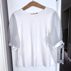 Vit topp med puffärmar i mesh - Superfin vit topp med transparenta puffärmar i mesh och rund halsringning. Ärmarna är korta och ger en cool, luftig känsla. Kroppen är i mjuk bomull och toppen har en enkel knapp i nacken. Perfekt för dig som gillar stilrena plagg med en twist.