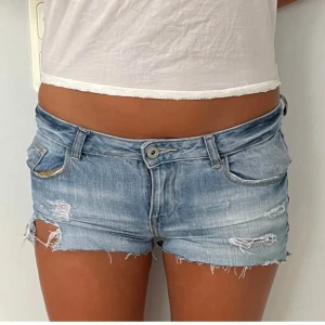 Lågmidjade ljusblå jeansshorts - Snygga lågmidjade jeansshorts i ljusblå denim med slitna detaljer och fransig kant. Klassisk femficksmodell med knapp och dragkedja framtill. Perfekta för varma dagar när du vill ha en avslappnad och trendig look.