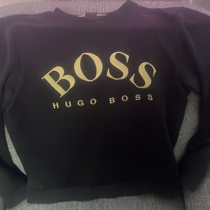 Svart sweatshirt från Hugo Boss - Svart sweatshirt från Hugo Boss med stort gult BOSS-tryck framtill. Klassisk rund halsringning och långa ärmar. Snygg och enkel design med gul detalj på insidan av kragen. Perfekt för dig som gillar stilrena märkesplagg.