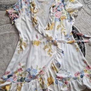 Blommig omlottklänning från ONLY - Supersöt kort omlottklänning från ONLY i vitt med blommigt mönster i pastelliga färger. Klänningen har volangkant, knyt i midjan och v-ringning. Perfekt för sommarens alla tillfällen och ger en romantisk vibe.