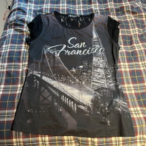 Svart spetsblus San Francisco Favor Fashion - Svart topp från Favor Fashion i storlek XL men sitter som s med genomskinlig spets och korta ärmar. Framsidan har ett tryck med San Francisco och stadssilhuett i svartvitt, medan baksidan är helspets med blommigt mönster. Perfekt för dig som gillar statement-plagg och coola detaljer.