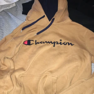 Beige Champion hoodie med broderad logga - Säljer en snygg beige hoodie från Champion med stor broderad logga i svart, rött och gult på bröstet. Tröjan har huva med mörkblå snören och insida, samt en klassisk känguruficka framtill. Perfekt för dig som gillar streetwear och vill ha en chill look.