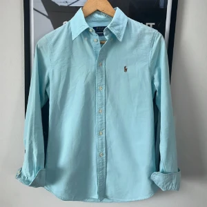 Ljusblå skjorta från Polo Ralph Lauren - Snygg ljusblå skjorta från Polo Ralph Lauren i bomull med klassisk krage och knappar framtill. Custom fit-modell med lång ärm och ikoniska broderade loggan på bröstet. Perfekt för dig som gillar stilrena och tidlösa plagg.
