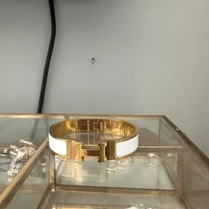 Hermès vit och guld Clic H armband - Snyggt Hermès Clic H armband i guldpläterad metall med vita emaljpaneler. Armbandet har den ikoniska H-formade låset framtill och är perfekt för att ge din outfit en lyxig touch. Enkel att öppna och stänga med vridlås.