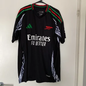 Arsenal Adidas svart matchtröja XL - Snygg svart Arsenal fotbollströja från Adidas i slim fit, storlek XL. Tröjan har gröna och röda ränder på axlarna, vit tryckt text och nummer 4 (White) på ryggen. Broderat rött kanonmärke på bröstet och coola mönsterdetaljer längs sidorna. Helt oanvänd så den är i nyskick även lappar sitter kvar. Storleken är XL men passar någon som är 180-185 cm lång. Vid minsta fundering hör av dig.