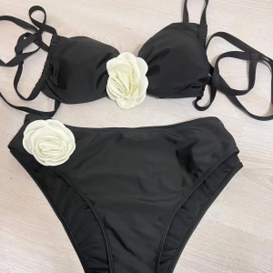 Svart bikini med vita blommor - Supersnygg svart bikini med stora vita blommor i tyg på både överdel och underdel. Bikinin har knytband och en klassisk trekantsform. 