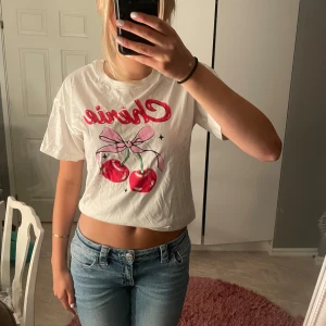 Vit croppad t-shirt med körsbär - Söt vit croppad t-shirt med tryck av röda körsbär och en rosa rosett framtill.🥰🥰storlek-170