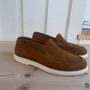 Bruna mockaloafers med vit sula - loafers i brun mocka från Loake, tillverkade i Portugal. Skorna har en klassisk slip-on design med vit platt sula och rund tå.  Nypris 2600kr.