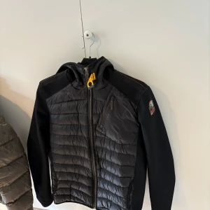 Svart pufferjacka från Parajumpers - Svart pufferjacka från Parajumpers med quiltad front, huva och dragkedja. Jackan har en gul detalj vid halsen och en bröstficka med dragkedja. Ärmarna är i ett stretchigt material och loggan sitter på vänster ärm. Ordinarie pris 5000