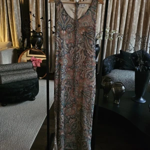 Paisleymönstrad långklänning från Dry Lake - Supersnygg långklänning från Dry Lake med paisleymönster i beige, blått, rött och grönt. Ärmlös modell med u-ringning och knytband tillkommer. Klänningen har slits i sidan och är tillverkad i ett lätt, glansigt material som känns lyxigt.