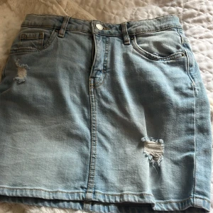 Ljusblå jeanskjol från LAB Industries - Snygg ljusblå jeanskjol från LAB Industries med slitna detaljer och klassiska fickor fram och bak. Kjolen har bälteshällor och stängs med knapp och dragkedja. Perfekt för dig som gillar en avslappnad och trendig stil.