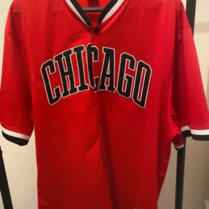 Röd baseballskjorta CHICAGO FSBN XL - Röd baseballinspirerad skjorta från FSBN i storlek XL med svartvita detaljer på krage och ärmslut. Stort CHICAGO-tryck framtill, knappar hela vägen och loose fit. Perfekt för dig som gillar sportig streetstyle.