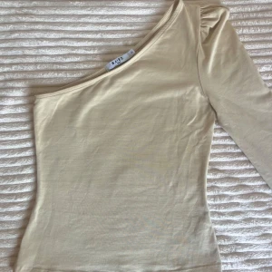 Beige one shoulder topp från NA-KD - Säljer en jättefin beige one shoulder topp från NA-KD med lång puffärm på ena sidan. Toppen är jättemjuk och skön, gjord utav 95% bomull. Jättefin topp som både är festlig men clean, passa på💗