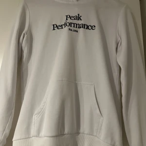 Vit hoodie Peak Performance - Vit hoodie från Peak Performance med svart broderad logga på bröstet. Klassisk känguruficka framtill, ribbade muddar och huva. Mjuk och skön, perfekt för chill dagar. Passar dig som gillar enkel och stilren look. Hoodien har lite slit men inget som märks eller påverkas. Den har även en liten missfärgning på baksidan av armen men syns nästan inte. 
